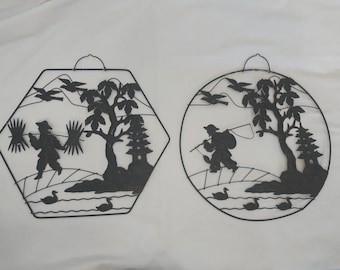 May include: Two black metal wall hangings with a Japanese theme. One is a hexagon shape with a tree, a pagoda, and a person carrying a bundle of sticks. The other is a round shape with a tree, a pagoda, and a person fishing.