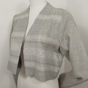 May include: A light gray, open-front jacket with long sleeves. The jacket features a woven pattern of white geometric designs across the chest and sleeves. The jacket is displayed on a mannequin.