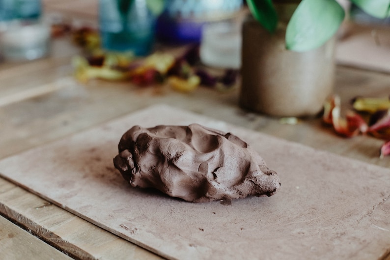 May include: A close-up of a brown clay lump on a light brown surface. The clay is soft and pliable, ready for sculpting.