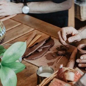 May include: A person is working with brown clay, shaping a small pot. The clay is on a wooden table with a wooden tool and a small piece of clay. There is a cup of milk and a plant in the background.