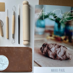 May include: A craft kit for making air dry clay projects. The kit includes a block of brown clay, a rolling pin, a small yellow sponge, a piece of sandpaper, a wooden tool, a metal tool, a small paintbrush, and a ring. The kit also includes a booklet with instructions and a logo that says "London Wildflowers".