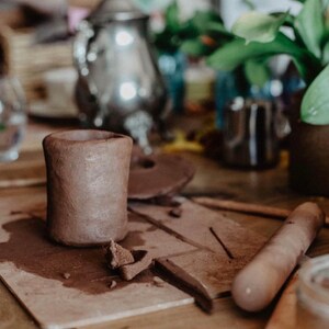 May include: A close-up of a brown clay mug on a table with a wooden rolling pin and a small bowl of clay. The mug is handmade and has a rustic look.