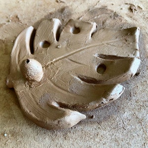 May include: A brown clay leaf with a hole in the centre and a small round knob on the side. The leaf has a textured surface and appears to be handmade.