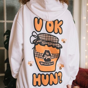 Pode incluir: Camisola de capuz branca com um gráfico de um pote de mel com o texto "U OK Hun?" e "Hey Hunny Spread the love" nele. O pote de mel é decorado com um padrão xadrez e há abelhas ao redor.