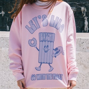 Peut inclure: Sweat-shirt rose avec un graphique d'un flotteur de piscine gonflable souriant tenant un beignet et le texte "Let's Lila Batchi Go With The Float".