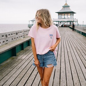 May include: A woman wearing a pink t-shirt with the text "SEA YA" on the front, blue denim shorts, and sunglasses. She is standing on a wooden pier with a gazebo in the background.
