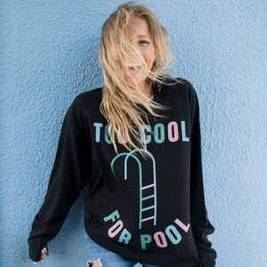 以下が含まれることがあります： ピンク、ブルー、グリーンの文字で「Too Cool For Pool」と書かれた黒いスウェットシャツ。テキストはプールラダーのグラフィックで囲まれています。