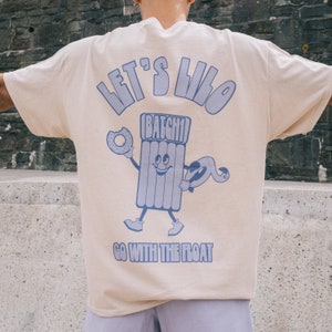 Peut inclure: Un t-shirt beige avec un motif bleu d'un personnage de dessin animé tenant un beignet et un flotteur de piscine. Le texte "Let's Lilo" et "Go with the Float" est imprimé sur le t-shirt.