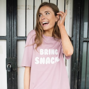 Puede incluir: Una mujer con una camiseta rosa con el texto "BRING SNACK" en letras blancas con forma de burbuja.