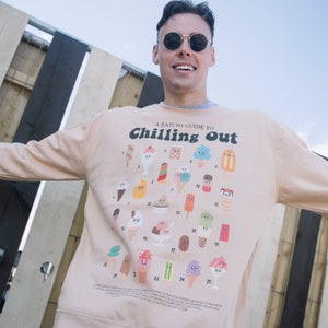 Op de afbeelding: Een licht beige sweatshirt met een grafische print van ijshoorntjes en ijslolly's. De tekst "A Batchi Guide to Chilling Out" is in zwart gedrukt op de voorkant van het sweatshirt.