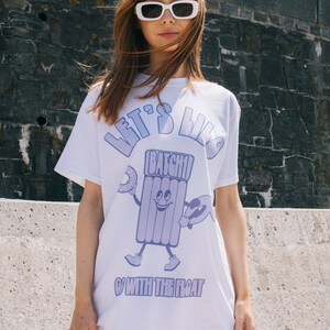 Peut inclure: T-shirt blanc avec un motif violet d'un flotteur de piscine de dessin animé tenant un beignet et une planche de surf. Le texte "Let's Live" et "Go With The Float" est imprimé sur le t-shirt.