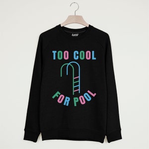 以下が含まれることがあります： カラフルな文字で「TOO COOL FOR POOL」と書かれた黒いスウェットシャツ。 「COOL」と「FOR」の間に、プールのはしごのスタイリッシュな画像があります。