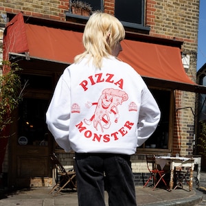 Op de afbeelding: Wit sweatshirt met een rode afbeelding van een pizzapunt met een gezicht en de tekst "Pizza Monster" op de rug.