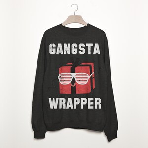 Könnte beinhalten: Dunkelgraues Sweatshirt mit dem weißen Schriftzug „GANSTA WRAPPER“. Eine rote Geschenkbox-Grafik mit weißer Sonnenbrille befindet sich in der Mitte. Das Sweatshirt hängt an einem Holzkleiderbügel.