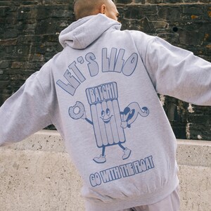 Peut inclure: Sweat-shirt à capuche gris avec un imprimé graphique bleu d'un personnage de dessin animé tenant un beignet et un flotteur de piscine. Le texte "Let's Lilo" et "Go with the float" est imprimé sur le sweat-shirt.