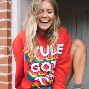 Peut inclure: Un sweat-shirt rouge avec un motif arc-en-ciel qui dit "Yule Got This". Le modèle porte le sweat-shirt et sourit.