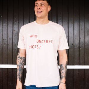 May include: A light pink t-shirt with the text "WHO ORDERED SHOTS?" printed in red.