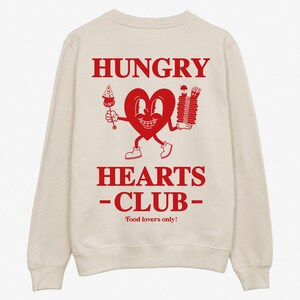 May include: A cream-colored sweatshirt with a red graphic print of a cartoon heart holding a slice of pizza and a stack of french fries. The text "HUNGRY HEARTS -CLUB- Food lovers only!" is printed below the graphic.