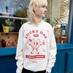 May include: Cream-colored sweatshirt with a red graphic of a smiling fortune cookie character holding a fortune and a box of cookies. The text "LUCKY DAY" and "FORTUNE COOKIES" are printed in red. The sweatshirt has long sleeves and a crew neck.