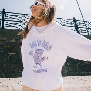 Peut inclure: Sweat-shirt blanc avec un personnage de dessin animé violet tenant un beignet et une planche de surf. Le texte "Let's Lilo with the Boat" est imprimé sur le sweat-shirt.