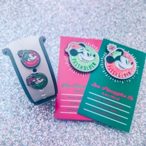 Puede incluir: Una pulsera MagicBand blanca y gris con dos pines de Mickey Mouse y dos tarjetas con pines de Mickey Mouse. Los pines presentan a Mickey Mouse con un gorro de Pap&aacute; Noel y el texto "Passholder". Las tarjetas son rosas y verdes.
