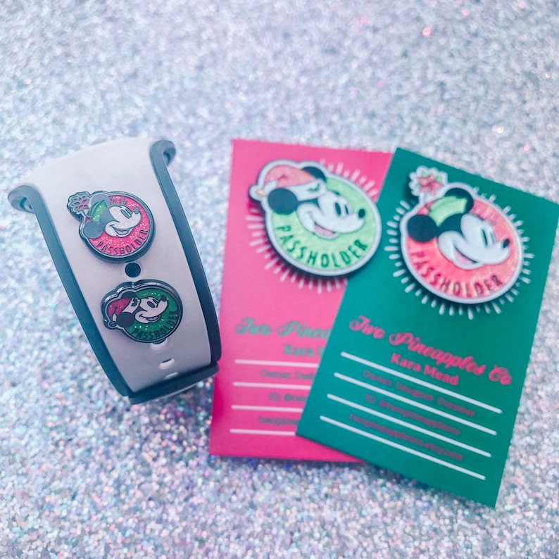 Puede incluir: Una pulsera MagicBand blanca y gris con dos pines de Mickey Mouse y dos tarjetas con pines de Mickey Mouse. Los pines presentan a Mickey Mouse con un gorro de Pap&aacute; Noel y el texto "Passholder". Las tarjetas son rosas y verdes.