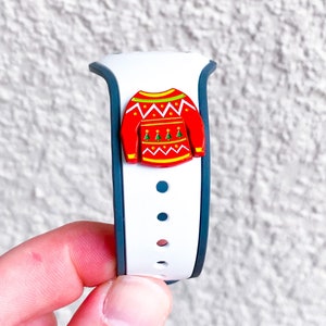 Könnte beinhalten: Weißes und blaues Silikonarmband mit einem roten und grünen Weihnachtspullover-Anhänger. Der Anhänger hat ein weißes Schneeflockenmuster und ein grünes Weihnachtsbaumdesign.