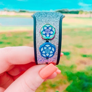 May include: A silver glitter Disney MagicBand with two blue and white Epcot park logo charms. The charms are iridescent and have a rainbow effect.