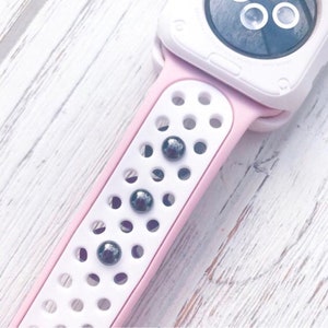 Puede incluir: Primer plano de una correa de reloj rosa y blanca con una esfera blanca. La correa tiene un dise&ntilde;o perforado con tres acentos circulares de color gris oscuro. La correa del reloj est&aacute; hecha de un material flexible.