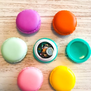 Puede incluir: Seis recipientes redondos de colores con forma de macarons. Los recipientes son de plástico y tienen un acabado liso y brillante. Un recipiente está abierto y revela una variedad de pequeñas pegatinas de colores en su interior.