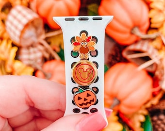 May include: A white Apple Watch band with three gold-colored charms. The charms are a turkey, a Mickey Mouse head, and a jack-o'-lantern. The band is surrounded by fall-colored pumpkins.
