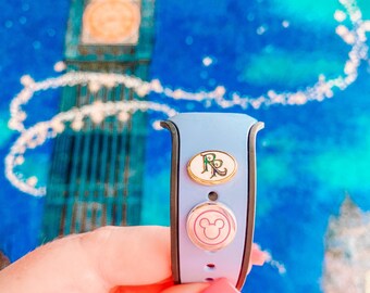 Puede incluir: Una pulsera MagicBand azul claro con dos pines decorativos. El pin superior es ovalado con un borde dorado y las letras "R" y "P". El pin inferior es redondo con una cabeza de Mickey Mouse. El fondo es una pintura azul y blanca del Big Ben.