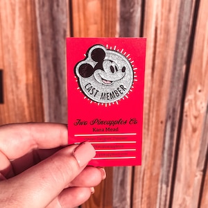 May include: Red business card with a silver glitter Mickey Mouse head with the text "Cast Member" inside a circle. The text "Two Pineapples Co. Kara Mead Owner, Designer, Dreamer" is below the Mickey Mouse head. The text "@twopineapplesco twopineapplesco.com" is at the bottom of the card.