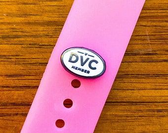 Puede incluir: Una correa de reloj rosa con un pin de miembro de DVC. El pin tiene forma ovalada con un fondo blanco y texto negro que dice "DVC MEMBER". La correa del reloj tiene varios agujeros circulares.
