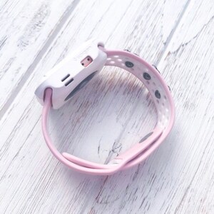 Puede incluir: Un reloj inteligente blanco y rosa con una correa rosa. El reloj tiene una esfera rectangular con una peque&ntilde;a pantalla y dos botones. La correa est&aacute; hecha de un material suave y flexible y tiene agujeros para ventilaci&oacute;n.