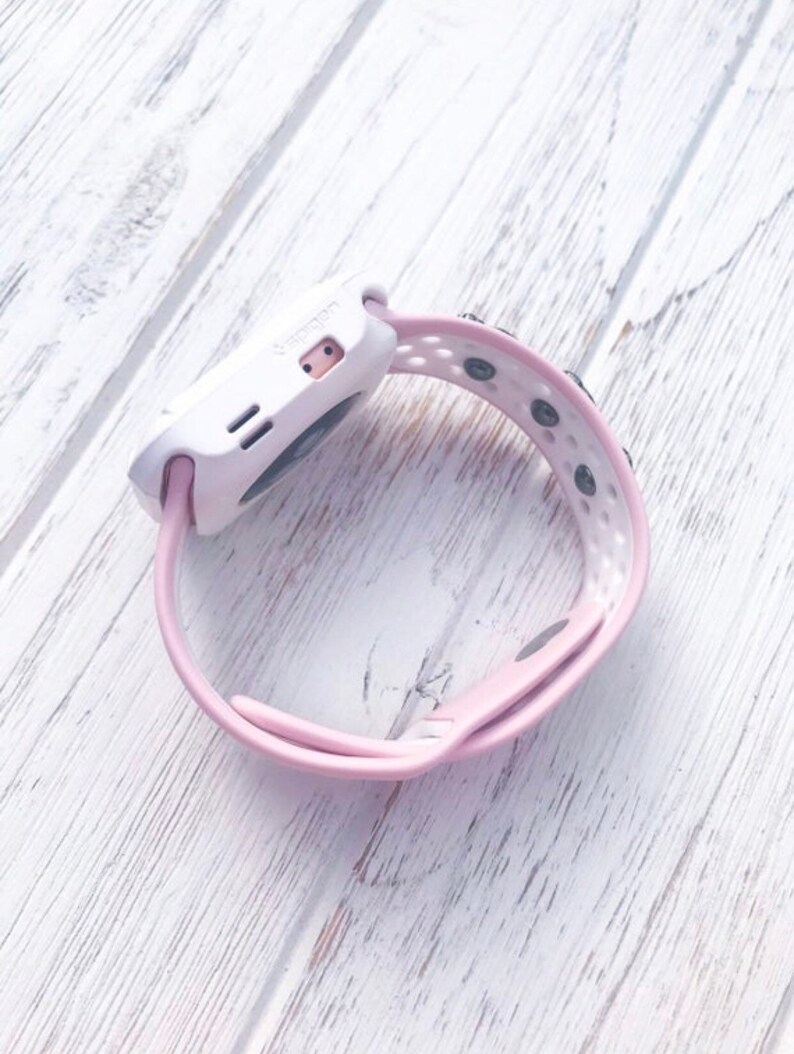 Puede incluir: Un reloj inteligente blanco y rosa con una correa rosa. El reloj tiene una esfera rectangular con una peque&ntilde;a pantalla y dos botones. La correa est&aacute; hecha de un material suave y flexible y tiene agujeros para ventilaci&oacute;n.