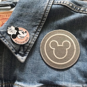 May include: A denim jacket with two embroidered patches. One patch features a cartoon image of Minnie Mouse with the text "Passholder". The other patch is a gray circle with a white outline of Mickey Mouse's ears.
