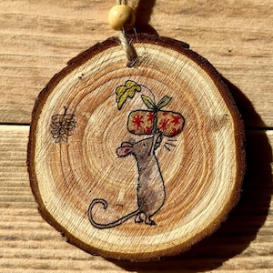 May include: A round wooden ornament featuring a painted image of a mouse carrying a wrapped gift. The mouse is brown and the gift is red and white with a yellow bow. The ornament has a twine hanger.