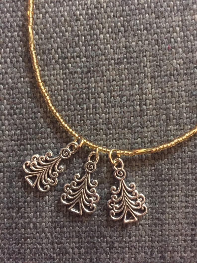 K&ouml;nnte beinhalten: Ein goldfarbenes Perlenarmband mit drei silbernen Anh&auml;nger in Form von stilisierten Weihnachtsb&auml;umen.