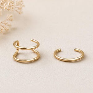 May include: Two gold-colored ear cuffs. One cuff has a double-band design, while the other is a single band. The jewelry is displayed on a neutral-toned surface, with dried flowers in the background.