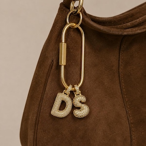 May include: A dark brown suede handbag featuring a gold-toned metal carabiner clip attached to the handle. Two gold-toned letter charms, 'D' and 'S', are suspended from the carabiner, both encrusted with small, clear, sparkling stones.