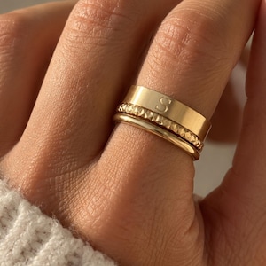 Può includere: Una pila di tre anelli color oro indossati su un dito. L'anello superiore è una fascia larga e liscia incisa con la lettera S. L'anello centrale presenta una superficie testurizzata e sfaccettata, mentre quello inferiore è un anello sottile e lucido.