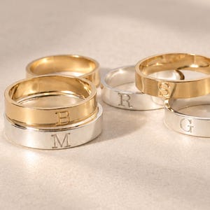 May include: A collection of five flat-band rings in gold and silver tones, each featuring a single engraved letter. The rings are arranged in a cluster, displaying the letters B, M, R, S, and G clearly on their smooth, polished metal surfaces.