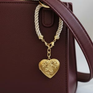 Può includere: Una borsa bordeaux con un ciondolo a forma di cuore dorato. Il medaglione presenta un motivo a rosa ed è appeso a una corda bianca e hardware dorato. La borsa ha una tracolla bordeaux abbinata.
