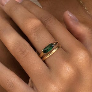 May include: A hand wearing two gold rings. The primary ring features a rectangular, emerald-green gemstone. The second ring is a textured gold band. A gold necklace is also visible.