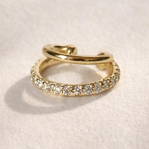 May include: A gold ear cuff featuring a double-band design. The lower band is encrusted with a row of small, clear, round-cut stones, while the upper band is a smooth, polished gold metal hoop. The piece has an open design for easy placement.