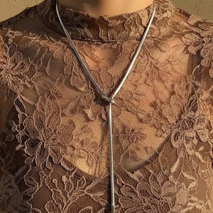 May include: A silver lariat necklace with a knotted design. The necklace hangs over a brown lace top with a high neckline. The lace pattern features floral designs.