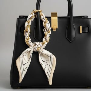 May include: Black leather handbag with a gold clasp and handle. A cream-colored scarf with a black and gold chain is looped around the handle. The scarf has a black line design and is tied in a knot.