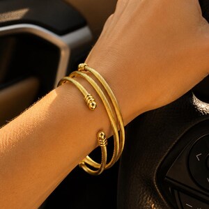 May include: A gold-toned cuff bracelet featuring a twisted cable texture and rounded ball ends. The bracelet is designed as a multi-wrap style, creating the appearance of three stacked bands around the wrist, with a polished metallic finish.