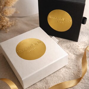 May include: Two jewelry boxes, one black and one white, with gold foil labels. The labels read "RuneandLight JEWELRY". A gold ribbon is draped across the surface. The boxes are closed, suggesting they contain jewelry.